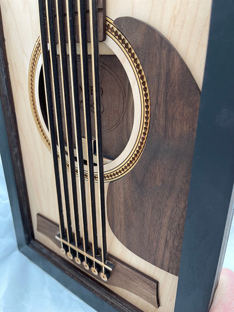 May include: A close-up of a wooden acoustic guitar with a black frame. The guitar has a sound hole with a decorative border and a wooden bridge with six tuning pegs.