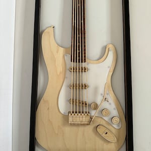 May include: A wooden model of an electric guitar in a black frame. The guitar is made of light wood and has a white pickguard. The guitar is displayed on a white wall.