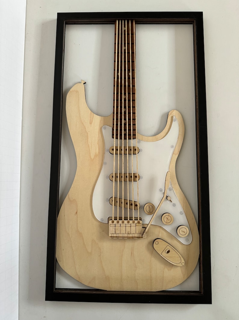May include: A wooden model of an electric guitar in a black frame. The guitar is made of light wood and has a white pickguard. The guitar is displayed on a white wall.