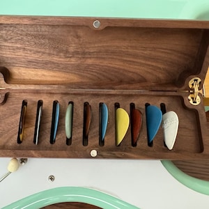 May include: A wooden guitar pick box with a hinged lid, open to reveal a row of ten colorful guitar picks.