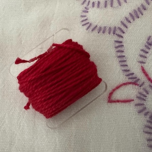 May include: A spool of bright red thread sits on a white fabric background. The thread is tightly wound on a clear plastic holder. In the background, there is an embroidered design with purple, pink, and yellow thread.