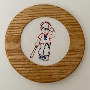 May include: Embroidered artwork of a young baseball player in a red and blue uniform, holding a bat and wearing a baseball cap. The artwork is set within a round, light-brown wooden frame.