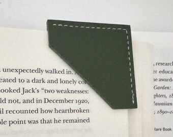 May include: A dark green leather bookmark with white stitching, placed on the corner of a book. The bookmark has a triangular shape and is made of a smooth material. The book's pages are visible.