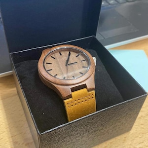 May include: A wooden wristwatch with a brown face and a tan leather band is displayed inside a black box. The watch has a minimalist design with simple hour markers and hands. The watch is a stylish accessory.