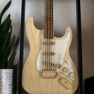May include: A wooden model of an electric guitar in a black frame. The guitar is made of light wood and has a white pickguard. The guitar is in a black frame with a glass front.