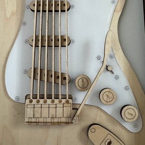 May include: Close-up of a wooden guitar body with a white pickguard. The guitar has a bridge, six strings, and three knobs labeled "Volume", "Inch", and "Tone".