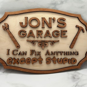 Puede incluir: Un letrero de madera con el texto "JON'S GARAGE I CAN FIX ANYTHING except STUPID" y una llave inglesa y un martillo a cada lado.