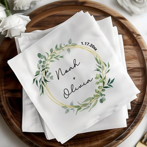 May include: White linen napkins with a gold and green wreath design. The wreath has the names Noah and Olivia and the date 1.17.20xx.