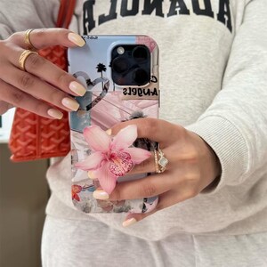 May include: A light blue and pink phone case with a floral design and the text "Los Angeles". The case features a heart-shaped cutout and a pink flower. The phone case is held by a person.