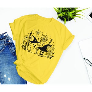 May include: A yellow t-shirt with a black and white graphic design of a ghost and a pumpkin wearing witch hats. The graphic is surrounded by black and white floral designs.