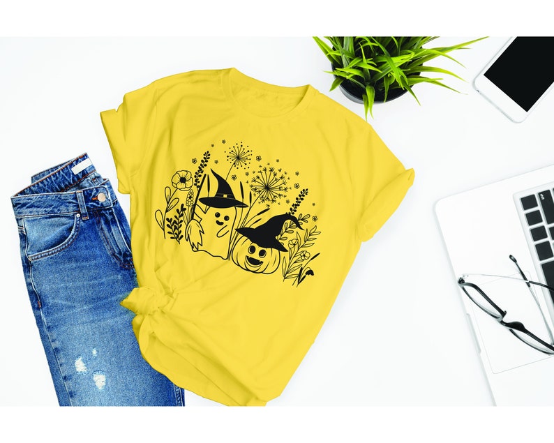 May include: A yellow t-shirt with a black and white graphic design of a ghost and a pumpkin wearing witch hats. The graphic is surrounded by black and white floral designs.