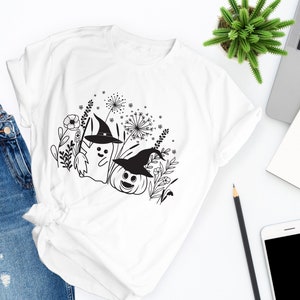 May include: White t-shirt with a black and white graphic design of a ghost and a pumpkin wearing witch hats in a field of flowers.
