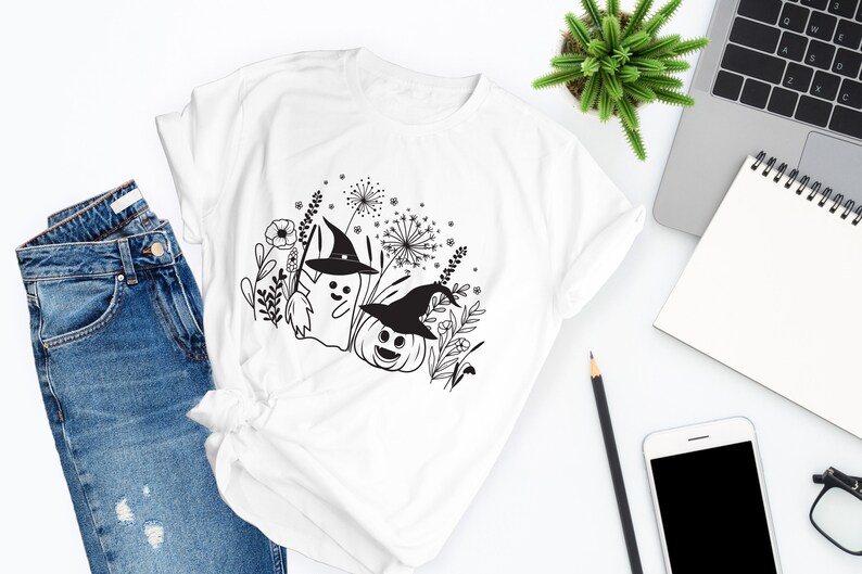 May include: White t-shirt with a black and white graphic design of a ghost and a pumpkin wearing witch hats in a field of flowers.