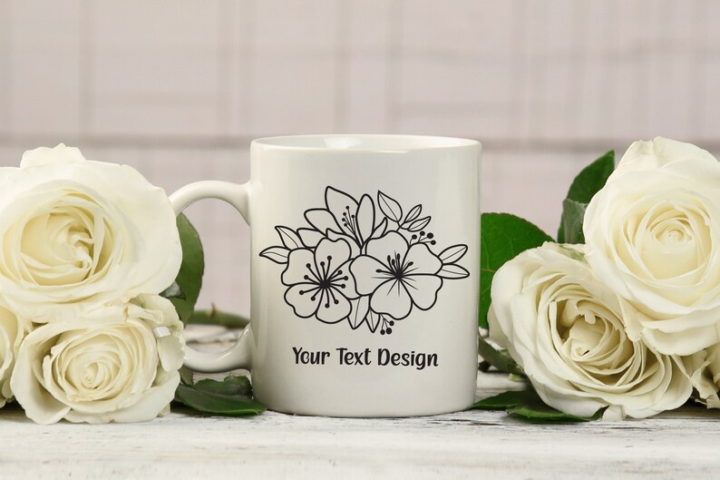 Pu&ograve; includere: Tazza in ceramica bianca con un design floreale nero e il testo "Your Text Design". La tazza &egrave; circondata da rose bianche.