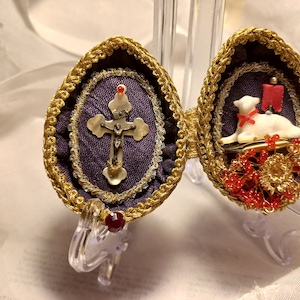 May include: Two ornate, gold-trimmed, egg-shaped containers with purple fabric interiors. One container features a silver crucifix with a pearl-like center, while the other contains a white porcelain lamb with a red ribbon and gold beads.