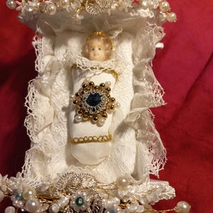 Könnte beinhalten: Eine weiße Baby Puppe in einem weißen Bettchen mit einer goldenen Krone und einem blauen Edelstein auf der Brust. Das Bettchen ist mit weißer Spitze und Perlen verziert.