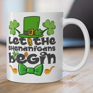 May include: White ceramic mug with a St. Patrick's Day design. The mug displays a green leprechaun hat, shamrocks, a green bow tie, and the words "Let the Shenanigans Begin" in black. A gold coin and pipes are also included.