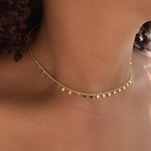 Può includere: Una collana a catena dorata con piccoli ciondoli rotondi. La collana è di tipo choker, che si adatta vicino al collo.