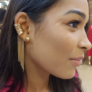 May include: A gold ear cuff and stud earring set. The ear cuff features multiple stacked rings that curve around the ear, with delicate chain tassels cascading down. The stud earring is a small, round gold ball. The jewelry is worn on the ear.