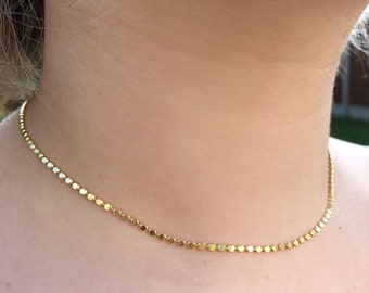 May include: A gold-toned necklace featuring a delicate chain composed of small, uniform, circular flat metal discs linked together in a continuous row. The necklace rests closely against the neck, showcasing a simple, reflective, and metallic design.
