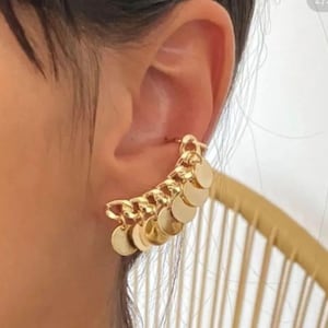 May include: A gold ear cuff with a chain design and multiple dangling circular charms. The jewellery piece is worn on the ear, highlighting its intricate details and reflective surface. The ear cuff is a fashionable accessory.
