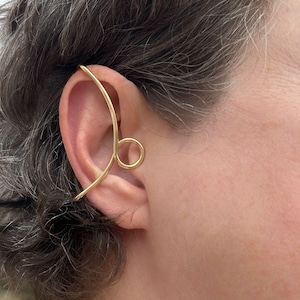 May include: A gold ear cuff is shown on an ear. The ear cuff has a curved design that wraps around the ear and features a circular element. The jewellery is made of a shiny, metallic material.
