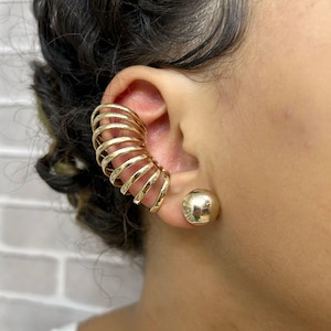 May include: Gold-colored ear cuff and stud earrings. The ear cuff features a spiral design that wraps around the ear. The stud earring is a simple, round ball shape. The jewelry is worn on a person's ear.