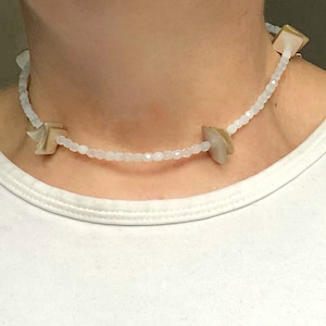 Puede incluir: Un collar tipo gargantilla con pequeñas cuentas blancas facetadas y translúcidas. A lo largo del hilo se intercalan varios componentes cuadrados de color blanco hueso iridiscente, similares a conchas, que añaden textura al diseño.