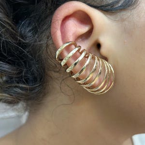 Könnte beinhalten: Goldfarbene Ohrklemme mit einem mehrschichtigen, spiralförmigen Design. Die Klemme umschließt das Ohr und erzeugt einen kühnen, modernen Look. Das Metall hat an einigen Stellen eine gehämmerten Textur.