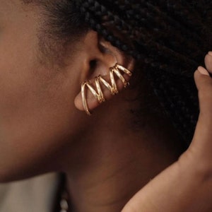May include: A gold-toned ear cuff featuring a continuous, multi-loop spiral design that wraps around the outer edge of the ear cartilage. The metal has a smooth, polished finish and a rounded, wire-like structure that creates a layered appearance.