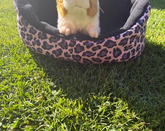 May include: A plush guinea pig toy with white and brown fur sitting inside a small, oval-shaped pet bed. The bed features a black interior lining and an exterior fabric pattern of purple and black leopard spots on a light pink background.