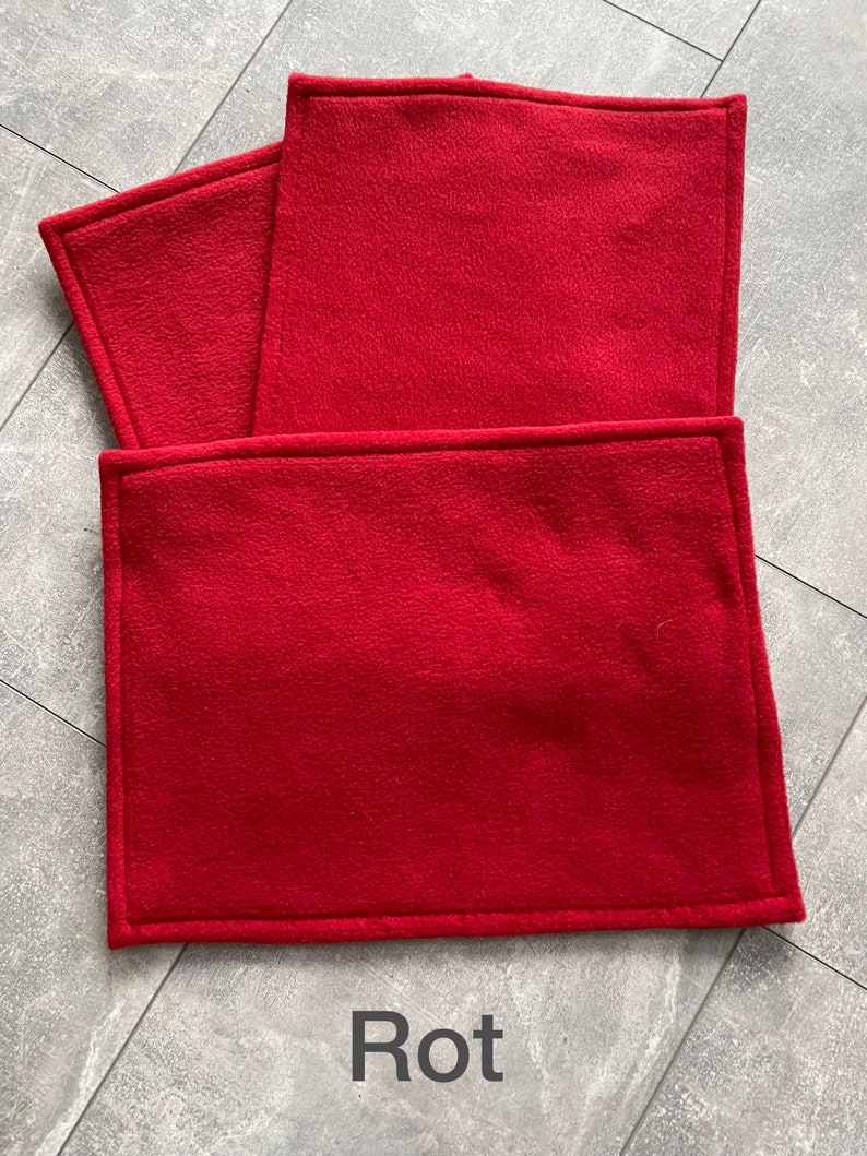 Peut inclure: Trois sets de table en polaire rouge avec un bord cousu. Les sets de table sont empil&eacute;s les uns sur les autres. Le mot "Rot" est &eacute;crit en noir sous les sets de table.