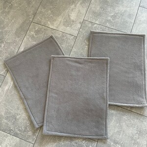 Peut inclure: Trois tapis rectangulaires en polaire gris clair avec un bord cousu. Les tapis sont sur un sol en carrelage gris. Le mot "Hellgrau" est &eacute;crit en noir sous les tapis.