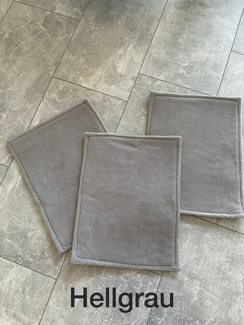 Peut inclure: Trois tapis rectangulaires en polaire gris clair avec un bord cousu. Les tapis sont sur un sol en carrelage gris. Le mot "Hellgrau" est &eacute;crit en noir sous les tapis.
