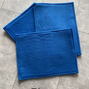 Peut inclure: Trois sets de table en polaire bleu avec un bord cousu. Les sets de table sont empil&eacute;s les uns sur les autres. Le mot "Blau" est &eacute;crit en noir sous les sets de table.