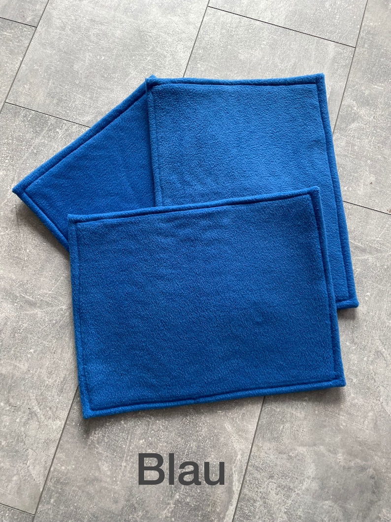 Peut inclure: Trois sets de table en polaire bleu avec un bord cousu. Les sets de table sont empil&eacute;s les uns sur les autres. Le mot "Blau" est &eacute;crit en noir sous les sets de table.