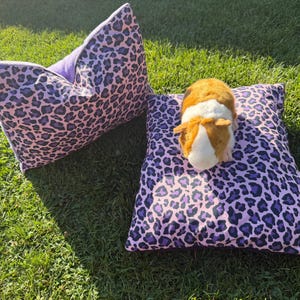 Peut inclure: Deux coussins rectangulaires avec un motif léopard violet et noir. Un coussin est placé à la verticale, tandis que l'autre est posé à plat avec un petit jouet en peluche représentant un cochon d'Inde marron et blanc au centre.