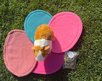May include: A plush guinea pig with brown and white fur sits atop four oval-shaped fleece mats in shades of teal, bright pink, and muted pink. Next to the mats is a small clear plastic bag containing dried herbs with a sticker that says THANK YOU.