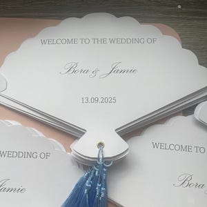 May include: A stack of white, shell-shaped paper wedding fans featuring the text WELCOME TO THE WEDDING OF, the names Bora & Jamie, and the date 13.09.2025 printed in gray. Each fan is secured with a gold-toned metal eyelet and a light blue tassel.
