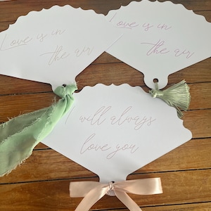 May include: Three white scalloped hand fans with thin script text. Two fans read Love is in the air, and one reads I will always love you. The fans are decorated with a sage green frayed ribbon, a sage green tassel, and a light pink satin ribbon.