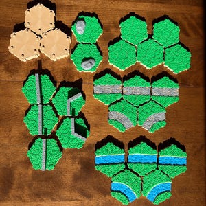 May include: Hexagonal terrain tiles in tan and green, arranged on a wooden surface. The green tiles depict grassy areas, roads, and rivers. The tan tiles appear to be unpainted. These tiles are likely for a tabletop game.