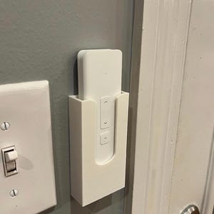 May include: A white remote control is held in a matching white wall-mounted holder. The remote has arrow and button symbols. The holder is mounted on a gray wall next to a white light switch. A white door is to the right.