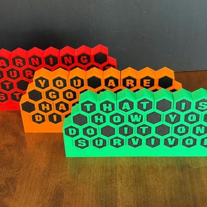 May include: Three colorful, honeycomb-shaped signs with motivational phrases. The signs are red, orange, and green, with black lettering. The red sign reads "EARNING YOUR AT ST", the orange sign reads "YOU GO HA DI ARE", and the green sign reads "THAT IS HOW YOU DO IT SURVIVOR".