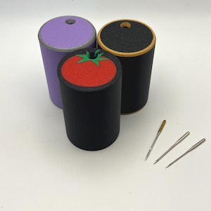 May include: Three cylindrical needle cases in purple, black, and black with a tomato design, and three needles. The cases have a small hole in the top. The needles are silver and gold. Sewing and craft supplies.