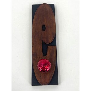 May include: A wooden number nine with a red, swirl-shaped embellishment in the center. The number is mounted on a black background.