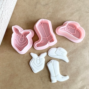 May include: Set of six pink and white plastic cookie cutters shaped like cowboy boots, hats, and bandanas.