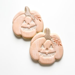 May include: Two light peach-colored pumpkin-shaped cookies decorated with a white face and a small sprig of leaves.