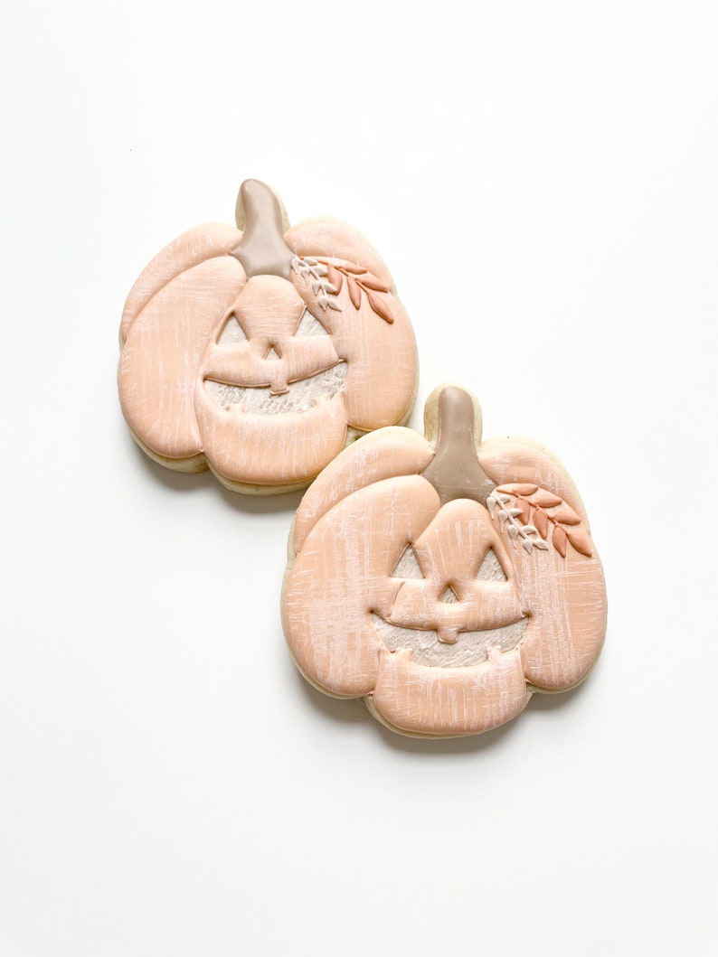May include: Two light peach-colored pumpkin-shaped cookies decorated with a white face and a small sprig of leaves.