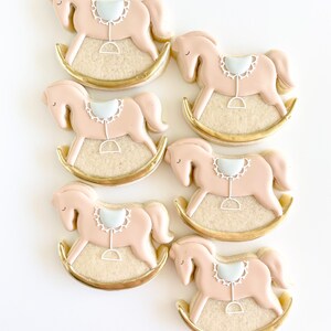 May include: Six pink and white rocking horse cookies decorated with gold icing. The cookies are arranged in a staggered pattern on a white surface.