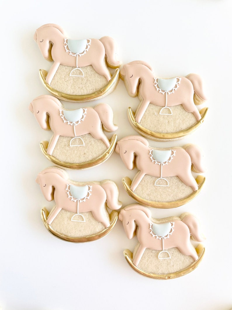May include: Six pink and white rocking horse cookies decorated with gold icing. The cookies are arranged in a staggered pattern on a white surface.